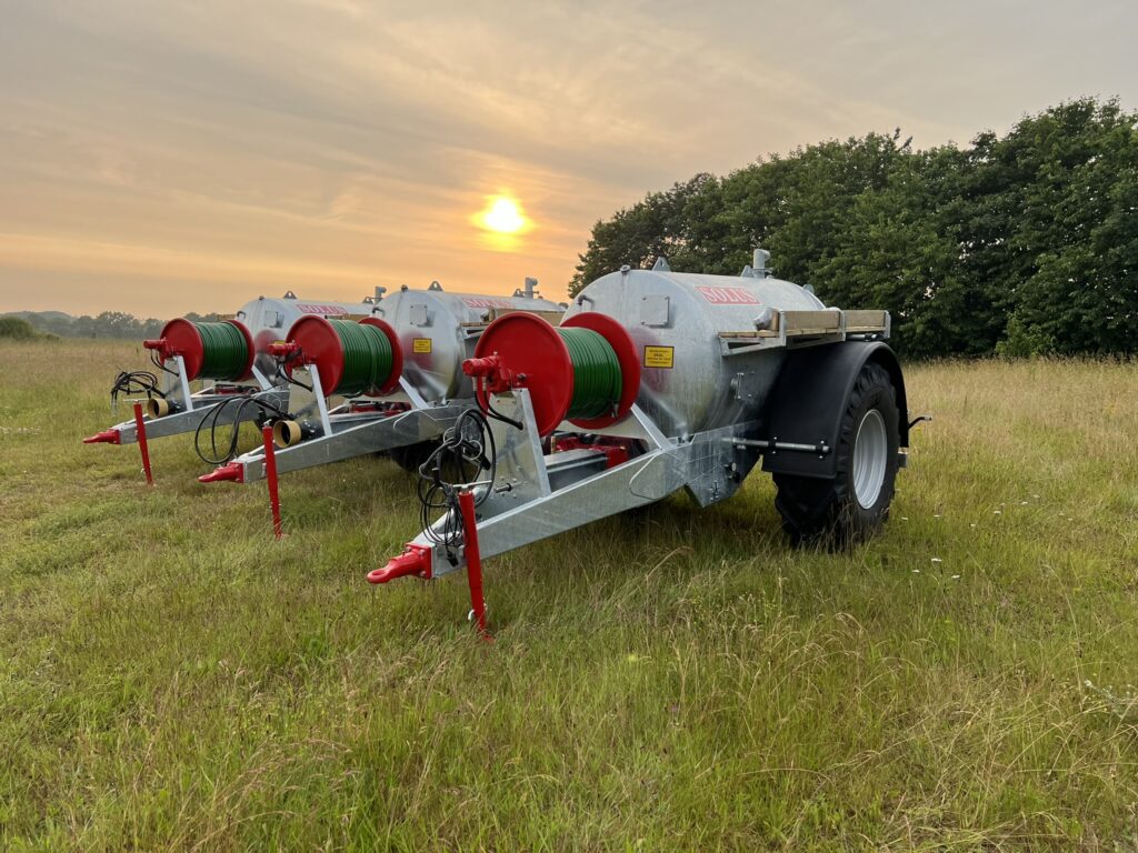 Tre landbrugsmaskiner placeret på en græsmark ved solnedgang. Maskinerne har røde og grønne detaljer og er udstyret med store hjul, der står klar til brug i det åbne landskab. I baggrunden ses træer og en smuk orange himmel.