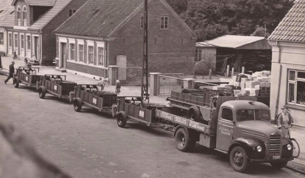 Sort-hvidt fotografi af en historisk lastbil med påhængsvogne fyldt med landbrugsudstyr, parkeret på en gade foran en række bygninger. Lastbilen har teksten 'Solus Kværne' og 'Tarm' malet på siden. I baggrunden ses flere bygninger og personer i arbejde, hvilket skaber en tidsbillede af industrielt arbejde i en landsby.